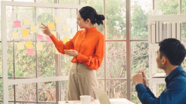 İş adamları ofisteki proje planlama kurulunda çalışıyor ve proje gelişimini analiz etmek için iş arkadaşlarıyla sohbet ediyorlar. Düzenli olması için cam duvara yapıştırılmış yapışkan notlar kullanıyorlar. .