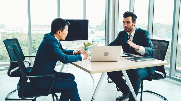 Two business people talk project strategy at office meeting room. Businessman discuss project planning with colleague at modern workplace while having conversation and advice on financial data report.