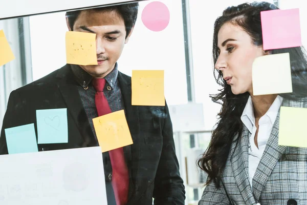 Business people work on project planning board in office and having conversation with coworker friend to analyze project development . They use sticky notes posted on glass wall to make it organized .