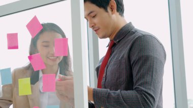 İş adamları ofisteki proje planlama kurulunda çalışıyor ve proje gelişimini analiz etmek için iş arkadaşlarıyla sohbet ediyorlar. Düzenli olması için cam duvara yapıştırılmış yapışkan notlar kullanıyorlar. .