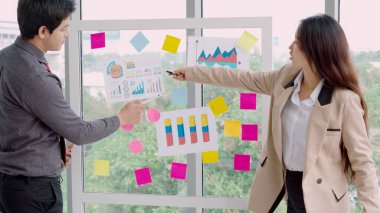 İş adamları ofisteki proje planlama kurulunda çalışıyor ve proje gelişimini analiz etmek için iş arkadaşlarıyla sohbet ediyorlar. Düzenli olması için cam duvara yapıştırılmış yapışkan notlar kullanıyorlar. .