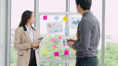İş adamları ofisteki proje planlama kurulunda çalışıyor ve proje gelişimini analiz etmek için iş arkadaşlarıyla sohbet ediyorlar. Düzenli olması için cam duvara yapıştırılmış yapışkan notlar kullanıyorlar. .