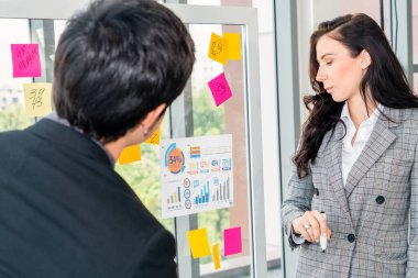 İş adamları ofisteki proje planlama kurulunda çalışıyor ve proje gelişimini analiz etmek için iş arkadaşlarıyla sohbet ediyorlar. Düzenli olması için cam duvara yapıştırılmış yapışkan notlar kullanıyorlar. .