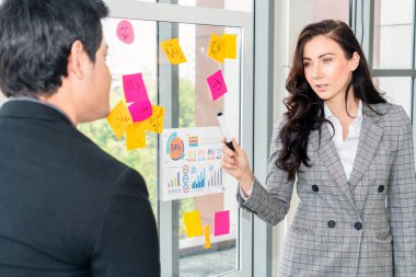 İş adamları ofisteki proje planlama kurulunda çalışıyor ve proje gelişimini analiz etmek için iş arkadaşlarıyla sohbet ediyorlar. Düzenli olması için cam duvara yapıştırılmış yapışkan notlar kullanıyorlar. .