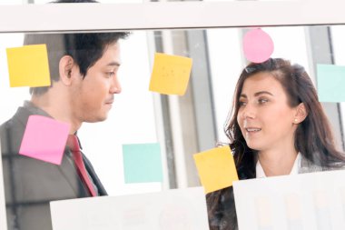 İş adamları ofisteki proje planlama kurulunda çalışıyor ve proje gelişimini analiz etmek için iş arkadaşlarıyla sohbet ediyorlar. Düzenli olması için cam duvara yapıştırılmış yapışkan notlar kullanıyorlar. .