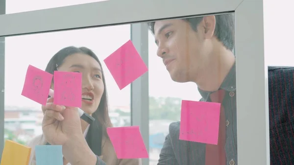 Business people work on project planning board in office and having conversation with coworker friend to analyze project development . They use sticky notes posted on glass wall to make it organized .