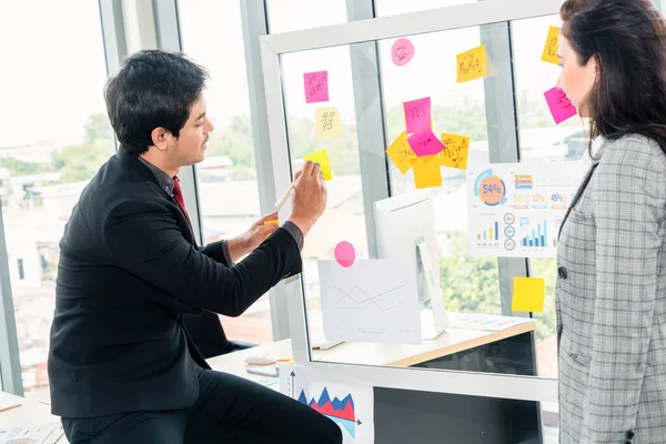 Business people work on project planning board in office and having conversation with coworker friend to analyze project development . They use sticky notes posted on glass wall to make it organized .