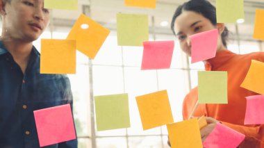 İş adamları ofisteki proje planlama kurulunda çalışıyor ve proje gelişimini analiz etmek için iş arkadaşlarıyla sohbet ediyorlar. Düzenli olması için cam duvara yapıştırılmış yapışkan notlar kullanıyorlar. .