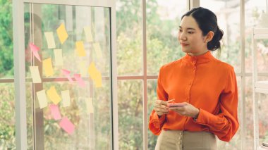 İş adamları ofisteki proje planlama kurulunda çalışıyor ve proje gelişimini analiz etmek için iş arkadaşlarıyla sohbet ediyorlar. Düzenli olması için cam duvara yapıştırılmış yapışkan notlar kullanıyorlar. .