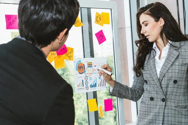 Business people work on project planning board in office and having conversation with coworker friend to analyze project development . They use sticky notes posted on glass wall to make it organized .