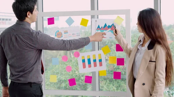 Business people work on project planning board in office and having conversation with coworker friend to analyze project development . They use sticky notes posted on glass wall to make it organized .