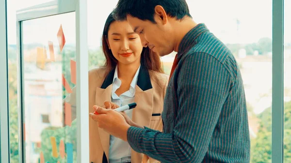 Business people work on project planning board in office and having conversation with coworker friend to analyze project development . They use sticky notes posted on glass wall to make it organized .