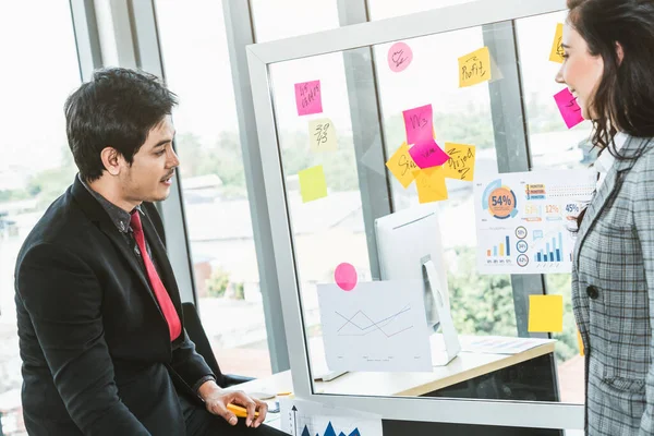 Business people work on project planning board in office and having conversation with coworker friend to analyze project development . They use sticky notes posted on glass wall to make it organized .