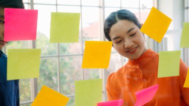 İş adamları ofisteki proje planlama kurulunda çalışıyor ve proje gelişimini analiz etmek için iş arkadaşlarıyla sohbet ediyorlar. Düzenli olması için cam duvara yapıştırılmış yapışkan notlar kullanıyorlar. .