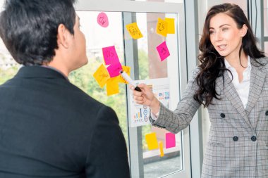 İş adamları ofisteki proje planlama kurulunda çalışıyor ve proje gelişimini analiz etmek için iş arkadaşlarıyla sohbet ediyorlar. Düzenli olması için cam duvara yapıştırılmış yapışkan notlar kullanıyorlar. .