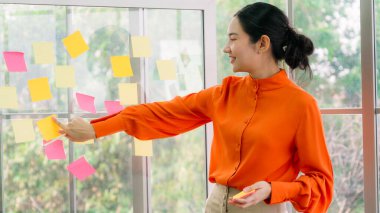 İş adamları ofisteki proje planlama kurulunda çalışıyor ve proje gelişimini analiz etmek için iş arkadaşlarıyla sohbet ediyorlar. Düzenli olması için cam duvara yapıştırılmış yapışkan notlar kullanıyorlar. .