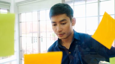 İş adamları ofisteki proje planlama kurulunda çalışıyor ve proje gelişimini analiz etmek için iş arkadaşlarıyla sohbet ediyorlar. Düzenli olması için cam duvara yapıştırılmış yapışkan notlar kullanıyorlar. .