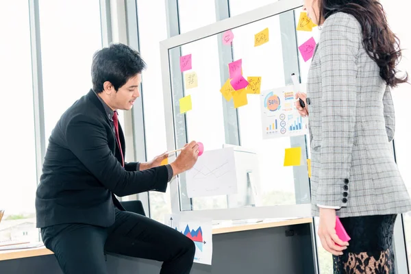 Business people work on project planning board in office and having conversation with coworker friend to analyze project development . They use sticky notes posted on glass wall to make it organized .