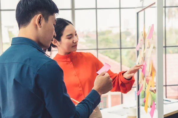 Business people work on project planning board in office and having conversation with coworker friend to analyze project development . They use sticky notes posted on glass wall to make it organized .