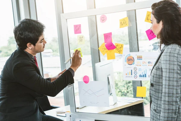 Business people work on project planning board in office and having conversation with coworker friend to analyze project development . They use sticky notes posted on glass wall to make it organized .