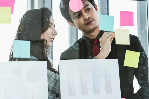 Business people work on project planning board in office and having conversation with coworker friend to analyze project development . They use sticky notes posted on glass wall to make it organized .