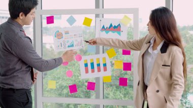 İş adamları ofisteki proje planlama kurulunda çalışıyor ve proje gelişimini analiz etmek için iş arkadaşlarıyla sohbet ediyorlar. Düzenli olması için cam duvara yapıştırılmış yapışkan notlar kullanıyorlar. .