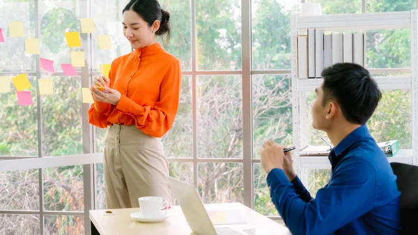 Business people work on project planning board in office and having conversation with coworker friend to analyze project development . They use sticky notes posted on glass wall to make it organized .