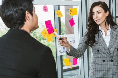 İş adamları ofisteki proje planlama kurulunda çalışıyor ve proje gelişimini analiz etmek için iş arkadaşlarıyla sohbet ediyorlar. Düzenli olması için cam duvara yapıştırılmış yapışkan notlar kullanıyorlar. .