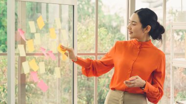 İş adamları ofisteki proje planlama kurulunda çalışıyor ve proje gelişimini analiz etmek için iş arkadaşlarıyla sohbet ediyorlar. Düzenli olması için cam duvara yapıştırılmış yapışkan notlar kullanıyorlar. .