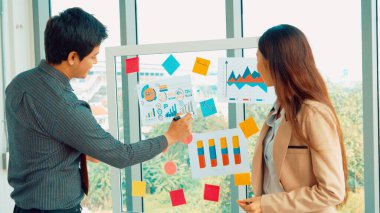İş adamları ofisteki proje planlama kurulunda çalışıyor ve proje gelişimini analiz etmek için iş arkadaşlarıyla sohbet ediyorlar. Düzenli olması için cam duvara yapıştırılmış yapışkan notlar kullanıyorlar. .