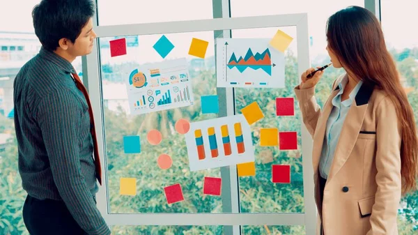 Business people work on project planning board in office and having conversation with coworker friend to analyze project development . They use sticky notes posted on glass wall to make it organized .