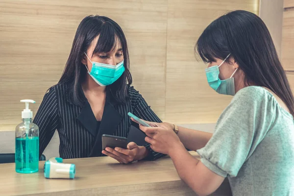 Receptionist and guest wearing face mask at front desk while having conversation in office or hospital . Covid 19 and coronavirus infection protection and protective policy concept .