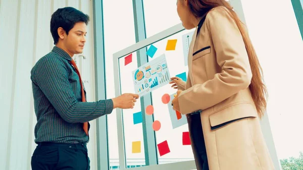 Business people work on project planning board in office and having conversation with coworker friend to analyze project development . They use sticky notes posted on glass wall to make it organized .