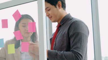 İş adamları ofisteki proje planlama kurulunda çalışıyor ve proje gelişimini analiz etmek için iş arkadaşlarıyla sohbet ediyorlar. Düzenli olması için cam duvara yapıştırılmış yapışkan notlar kullanıyorlar. .