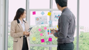 İş adamları ofisteki proje planlama kurulunda çalışıyor ve proje gelişimini analiz etmek için iş arkadaşlarıyla sohbet ediyorlar. Düzenli olması için cam duvara yapıştırılmış yapışkan notlar kullanıyorlar. .