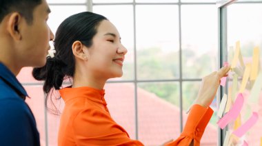 İş adamları ofisteki proje planlama kurulunda çalışıyor ve proje gelişimini analiz etmek için iş arkadaşlarıyla sohbet ediyorlar. Düzenli olması için cam duvara yapıştırılmış yapışkan notlar kullanıyorlar. .