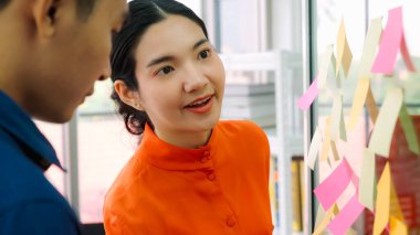 İş adamları ofisteki proje planlama kurulunda çalışıyor ve proje gelişimini analiz etmek için iş arkadaşlarıyla sohbet ediyorlar. Düzenli olması için cam duvara yapıştırılmış yapışkan notlar kullanıyorlar. .