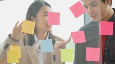 İş adamları ofisteki proje planlama kurulunda çalışıyor ve proje gelişimini analiz etmek için iş arkadaşlarıyla sohbet ediyorlar. Düzenli olması için cam duvara yapıştırılmış yapışkan notlar kullanıyorlar. .