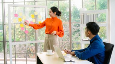 İş adamları ofisteki proje planlama kurulunda çalışıyor ve proje gelişimini analiz etmek için iş arkadaşlarıyla sohbet ediyorlar. Düzenli olması için cam duvara yapıştırılmış yapışkan notlar kullanıyorlar. .