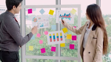 İş adamları ofisteki proje planlama kurulunda çalışıyor ve proje gelişimini analiz etmek için iş arkadaşlarıyla sohbet ediyorlar. Düzenli olması için cam duvara yapıştırılmış yapışkan notlar kullanıyorlar. .