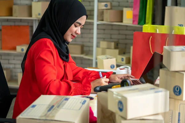 Muslim online seller at home office with shipping box for delivery to customer. Small business owner or entrepreneur doing e-commerce business on the internet.