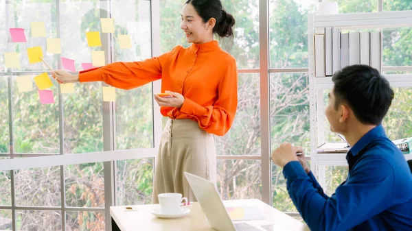 İş adamları ofisteki proje planlama kurulunda çalışıyor ve proje gelişimini analiz etmek için iş arkadaşlarıyla sohbet ediyorlar. Düzenli olması için cam duvara yapıştırılmış yapışkan notlar kullanıyorlar. .