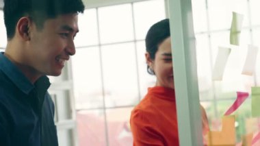 İş adamları ofisteki proje planlama kurulunda çalışıyor ve proje gelişimini analiz etmek için iş arkadaşlarıyla sohbet ediyorlar. Düzenli olması için cam duvara yapıştırılmış yapışkan notlar kullanıyorlar. .