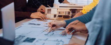 Çeşitli bir ekip beyin fırtınası yapıyor ve mimari bir proje planlıyor. Olay yeri, yaratıcı bir çalışma ortamında modeller ve çizimler üzerinde çalışan elleri gösteriyor. SACTR