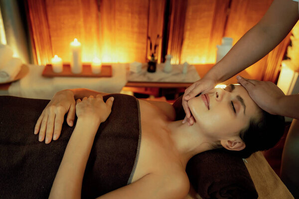 Caucasian woman enjoying relaxing anti-stress head massage and pampering facial beauty skin recreation leisure in warm candle lighting ambient salon spa in luxury resort or hotel. Quiescent