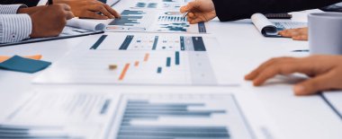 Bir grup profesyonel, kağıt üzerinde sergilenen tablo ve grafikleri analiz ederek modern bir ofis ortamında takım çalışması ve stratejik planlamayı vurguluyor. SACTR