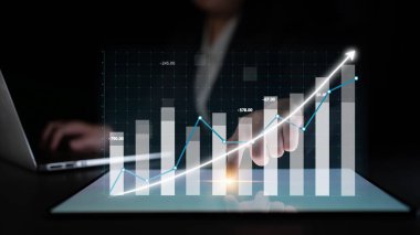 A hand analyzes a digital chart on a tablet, showcasing business growth through statistics and trends with a laptop in a modern workspace. Impute