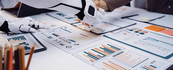Robot bir kol, modern bir ofiste işbirliğini sembolize eden bir insanla tokalaşır. Takım çalışmaları chartlar tarafından temsil edilen veri odaklı analizler üzerine yoğunlaşır. Noogenesis