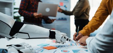 İşbirlikçi bir ortamda dokümanlar, dizüstü bilgisayarlar ve veri analizlerini tartışan profesyonellerle uğraşan bir robot asistanını gösteren modern bir ofis sahnesi. Noogenesis