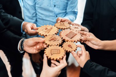 Hands from diverse individuals come together to assemble wooden gears featuring various icons, symbolizing teamwork and collaboration in a business environment. Amity